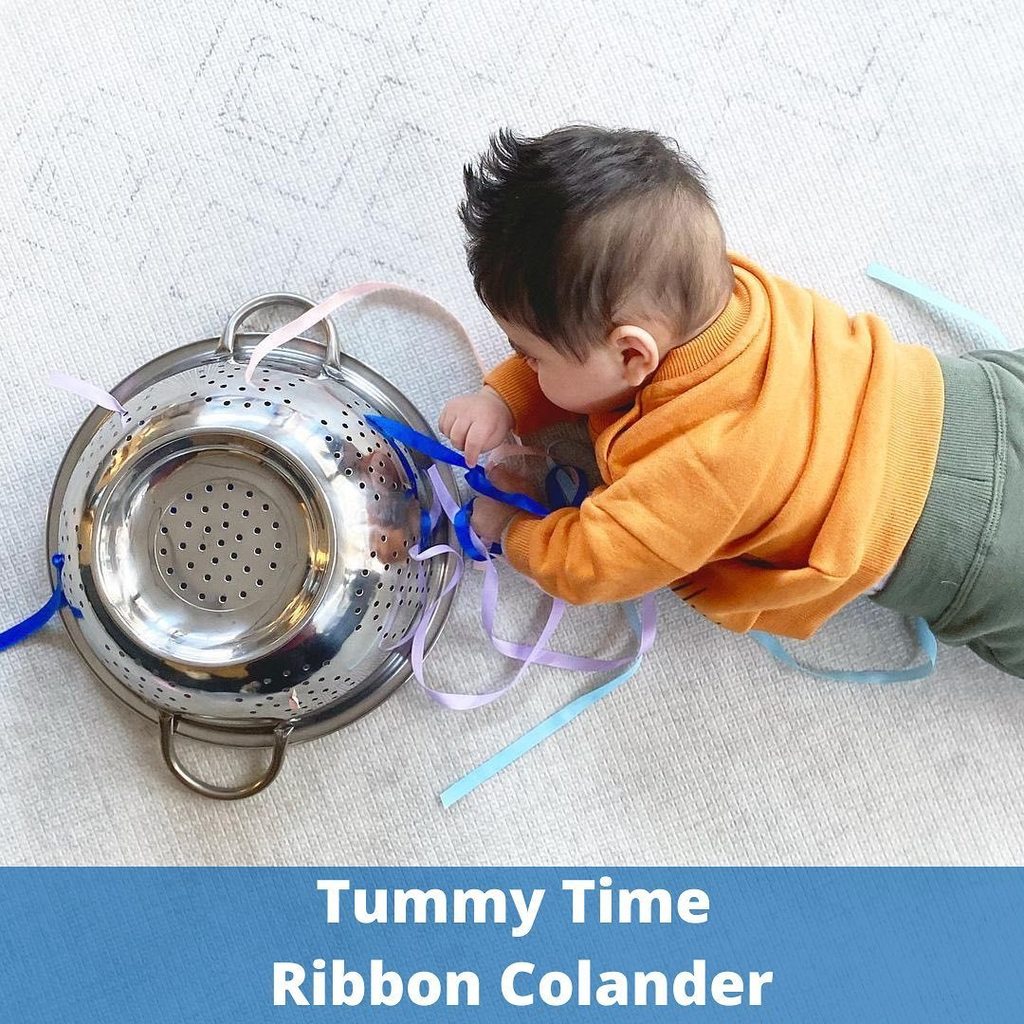 Baby on tummy playing beside colander with ribbons tied to it