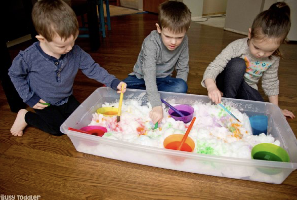 3 kids playing in snow that is in a tote, inside, with different colors of water paint