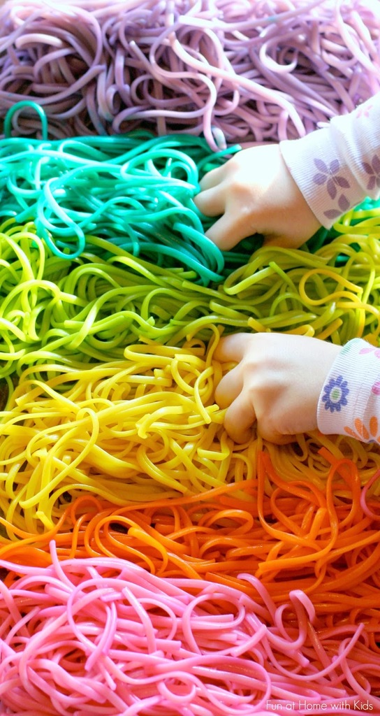 Shows a child's hands touching spaghetti noodles that have been dyed pink, orange, green, blue, and purple.